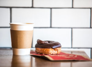 Coffee in a paper cup sits next to a flour donut with chocolate frosting on top.