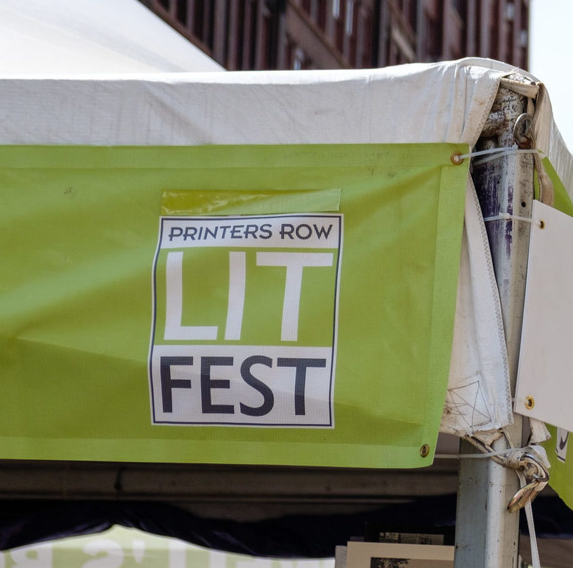 A foreground shot of a promotional sign for the Printers Row Lit Fest is in front of a giant red brick clock tower in the background